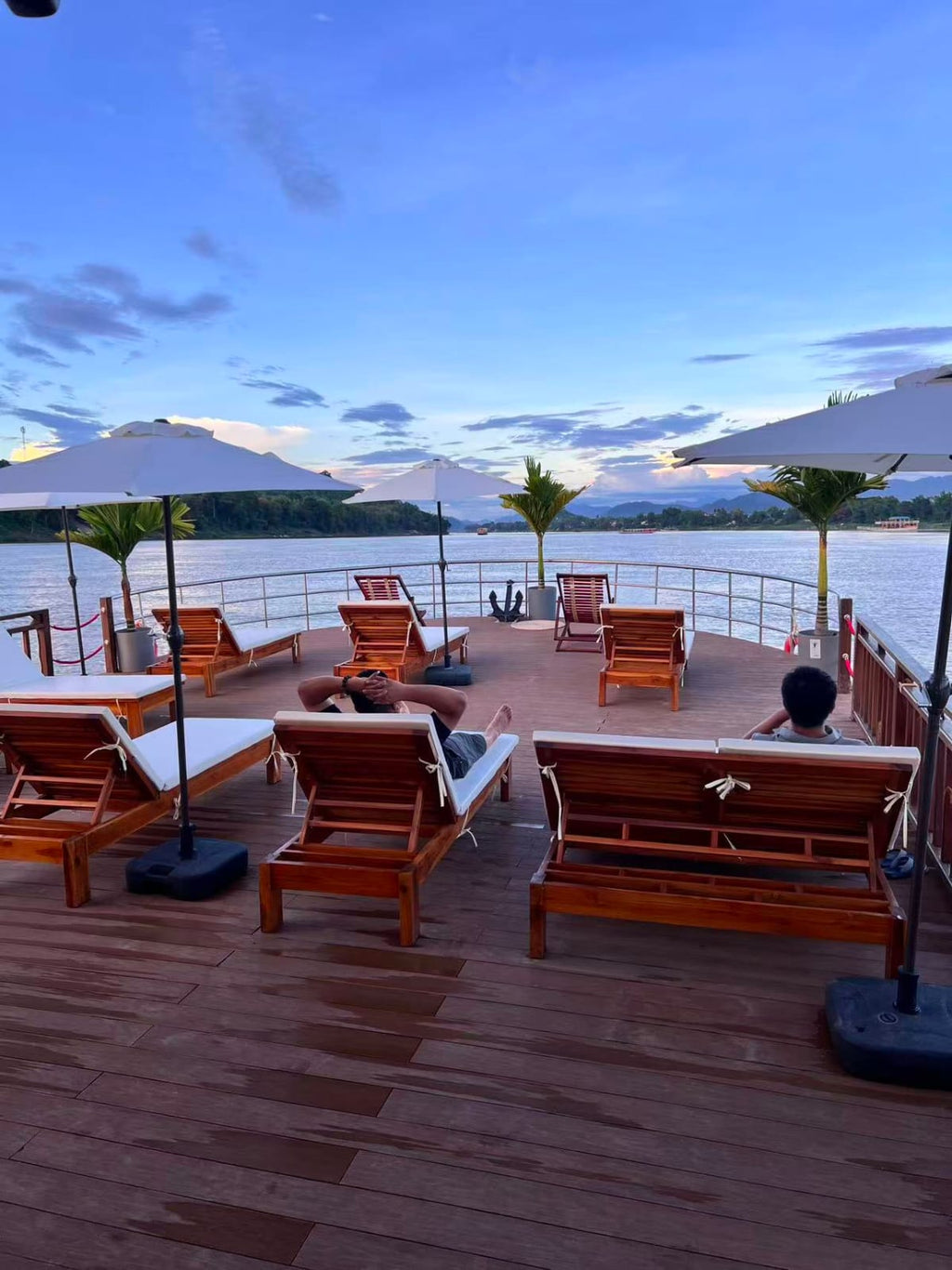 Wooden deck with lounge chairs and umbrellas overlooking a body of water with mountains in the background.